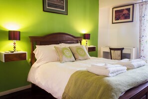 Un lit bien fait, avec du linge de lit blanc et des coussins verts, deux lampes de chevet et un tableau encadré au-dessus du lit.