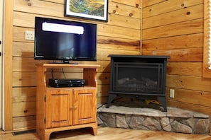 A wooden cabin interior with a television on a wooden stand, a stone fireplace, and a framed picture on the wall. - Rustic Cabin Rental near the Gauley River in West Virginia (Hico)