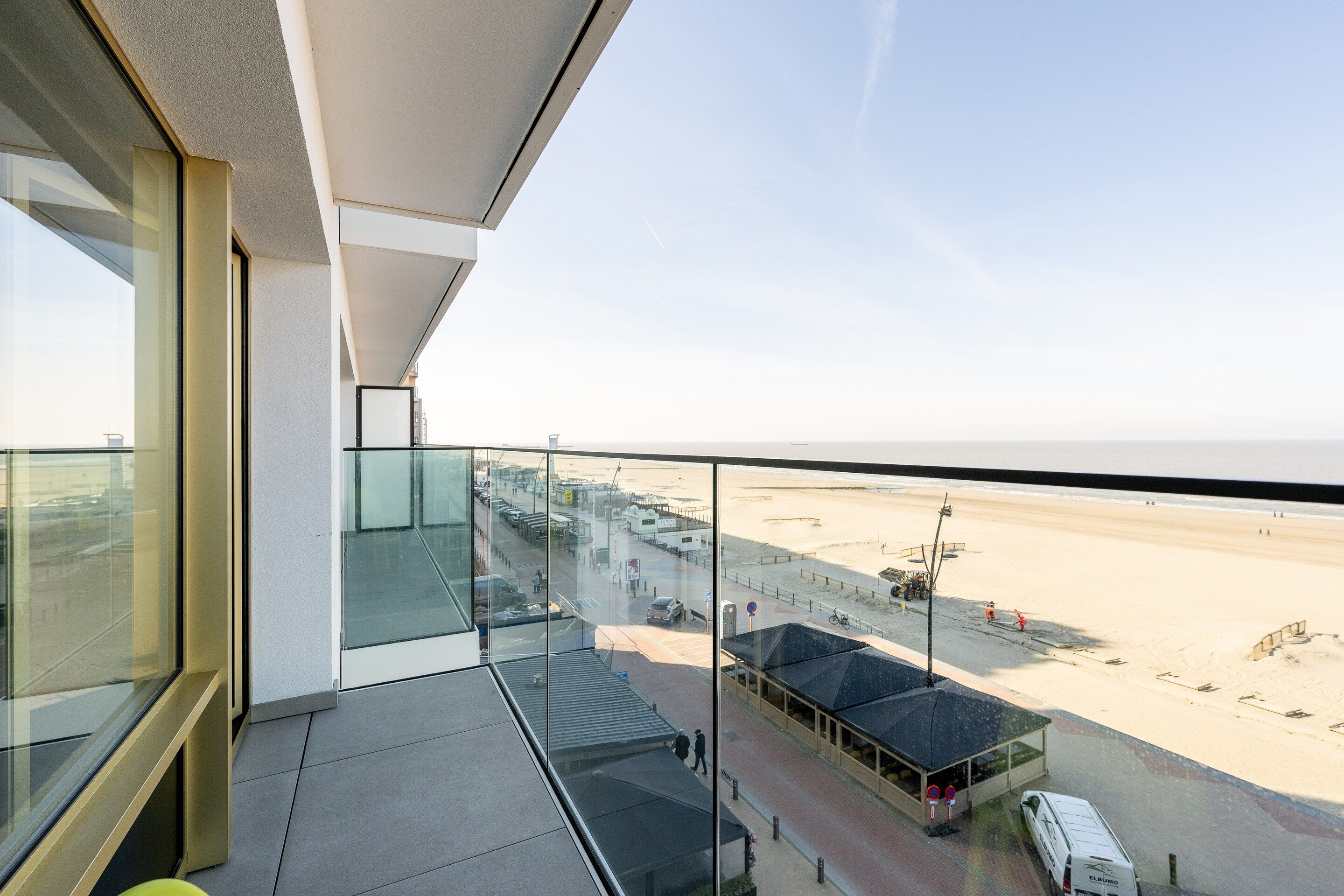 A balcony with a glass railing overlooking a beach and a parking area.