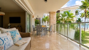 A balcony with a dining table and chairs, a sofa with decorative pillows, and a view of palm trees and a building. - The Ocean Is Your Window: 2-Bedroom Apartment at Playa Palmera (Punta Cana)