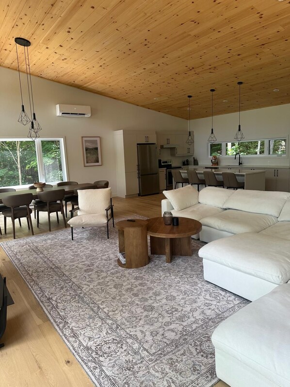 Un salón amplio con un sofá blanco grande, una mesa de comedor con sillas, una zona de cocina y vistas al exterior a través de ventanas grandes.