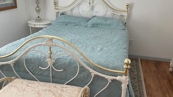 A bed with a white metal frame and a light blue bedspread, a bedside table with a lamp, and a patterned rug on the floor.