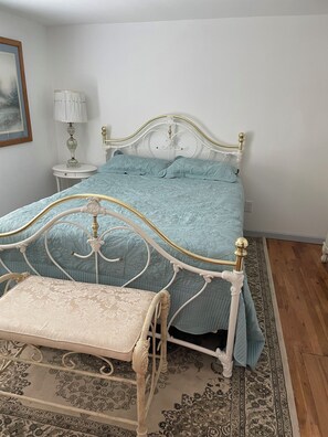 A bed with a white metal frame and a light blue bedspread, a bedside table with a lamp, and a patterned rug on the floor.