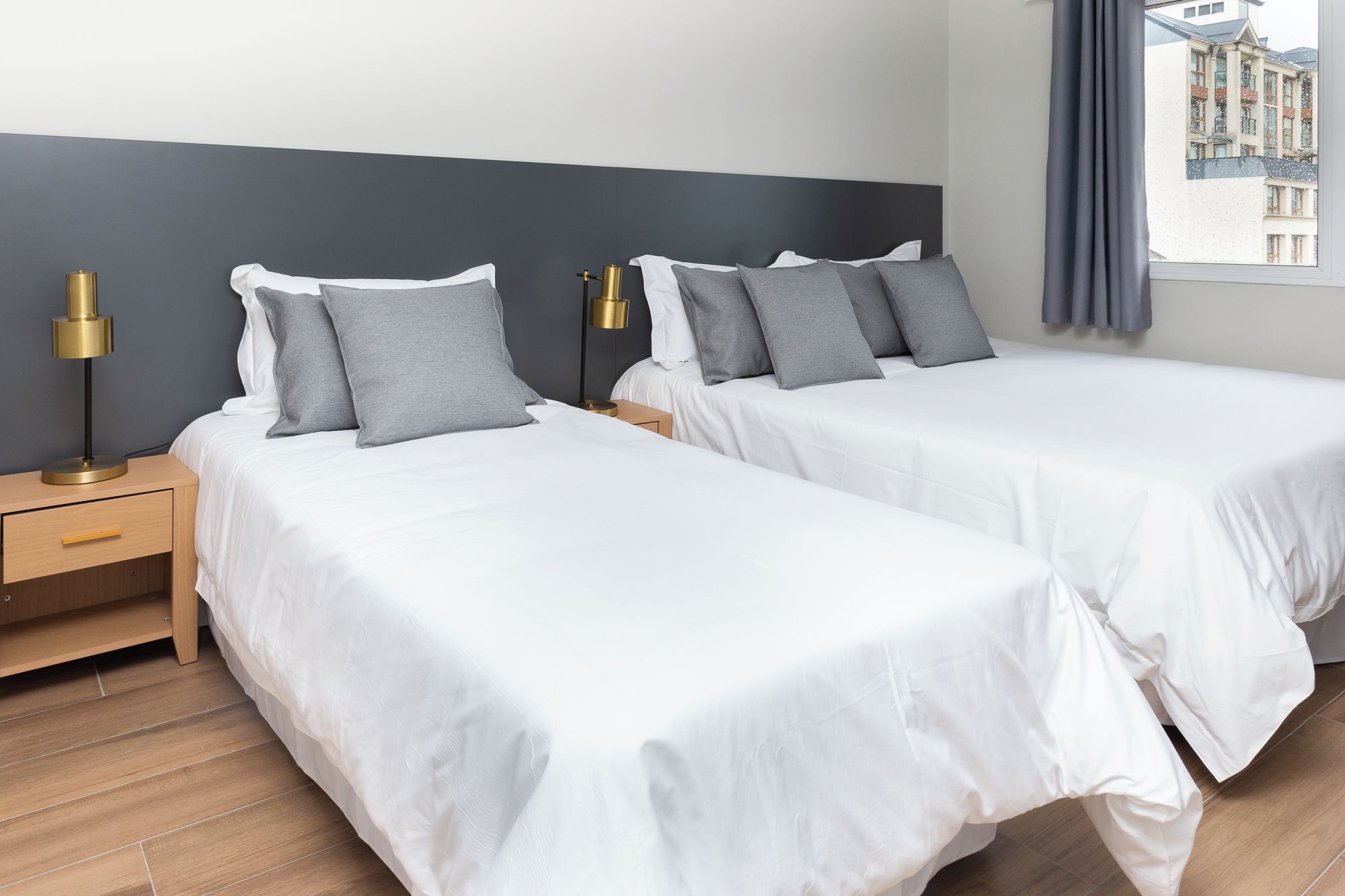 Quarto de hotel com duas camas, uma mesa de cabeceira, uma luminária e uma janela com cortinas.