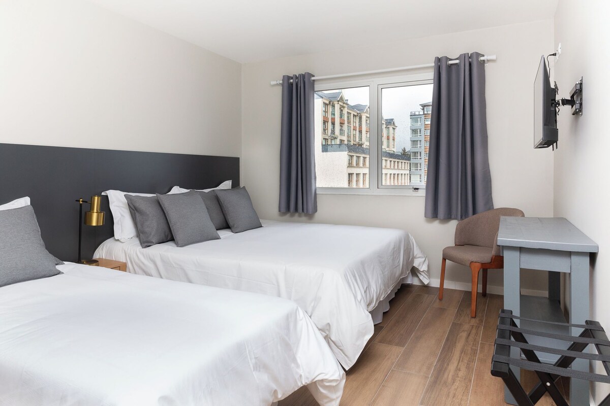 Una habitación de hotel con dos camas, un escritorio, una silla y un ventanal con cortinas.