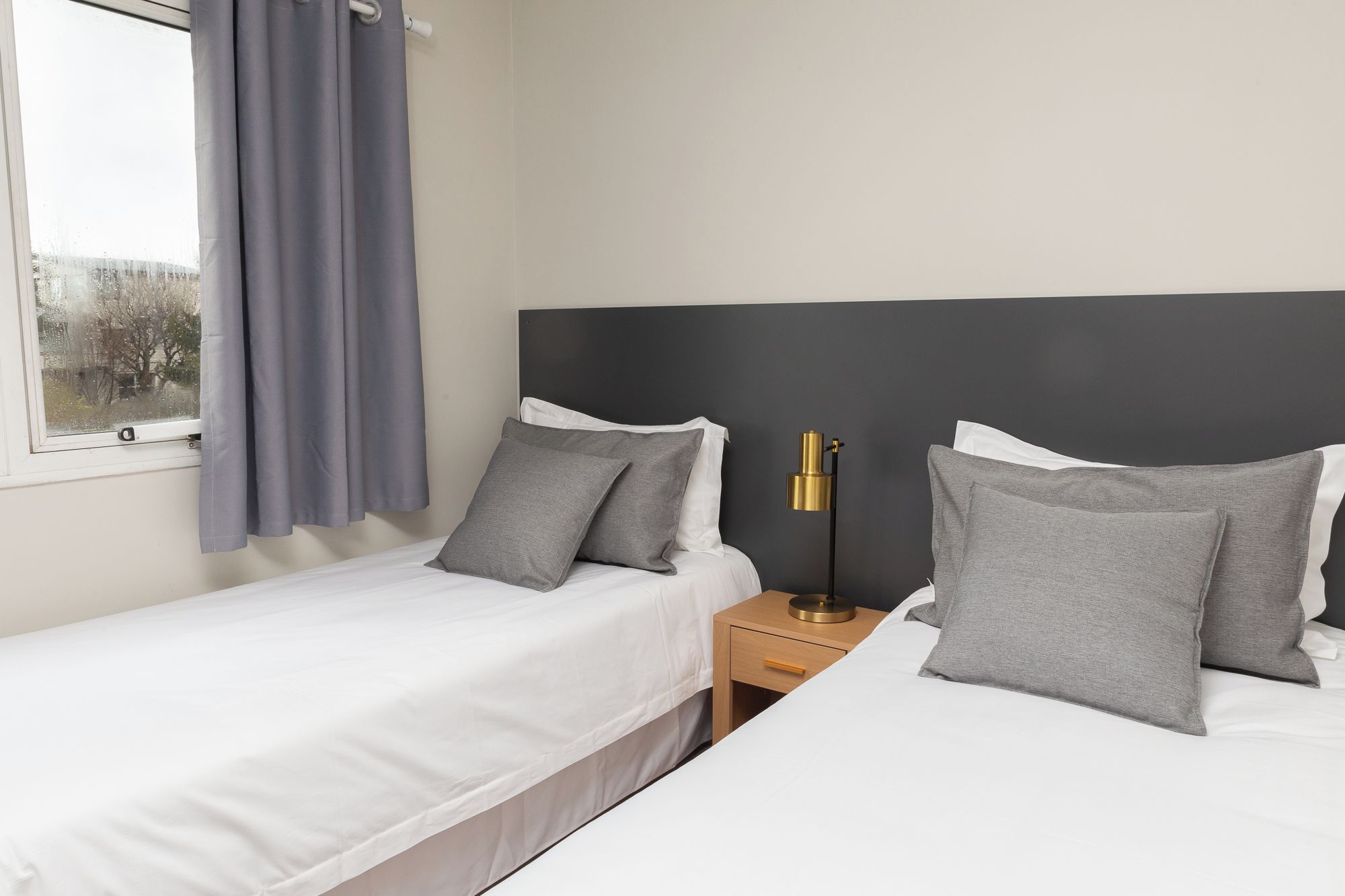 Une chambre d’hôtel avec deux lits simples, une table de chevet avec une lampe et une fenêtre avec des rideaux.
