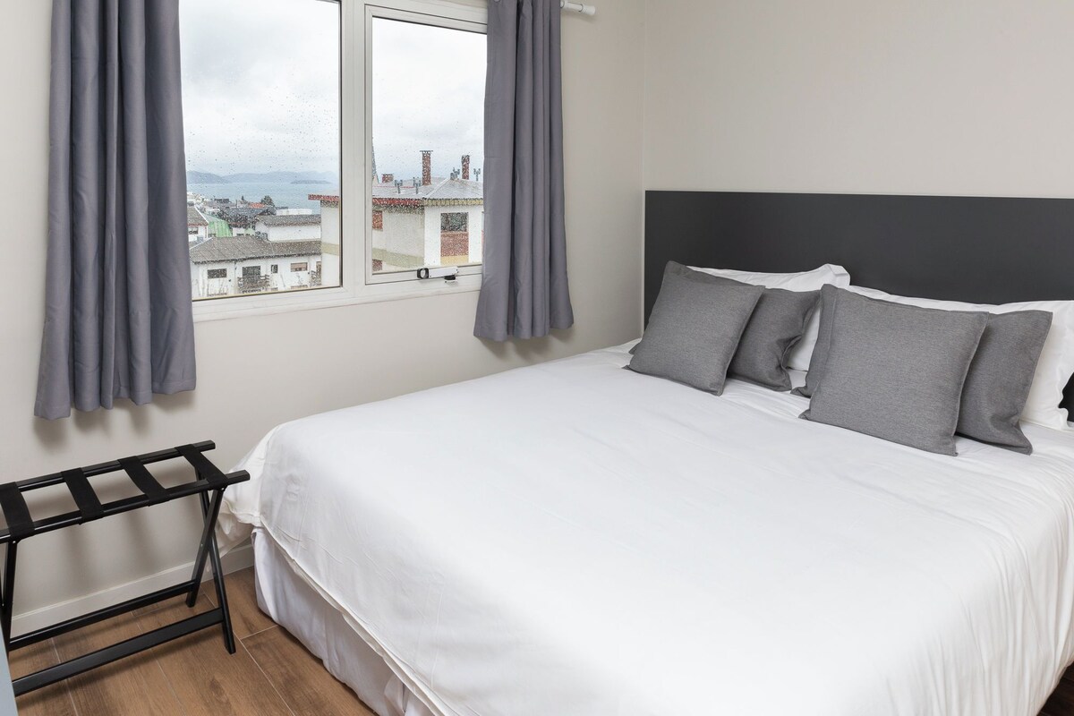Una cama bien hecha con ropa de cama blanca y almohadas grises, una ventana con cortinas grises y vistas a edificios.