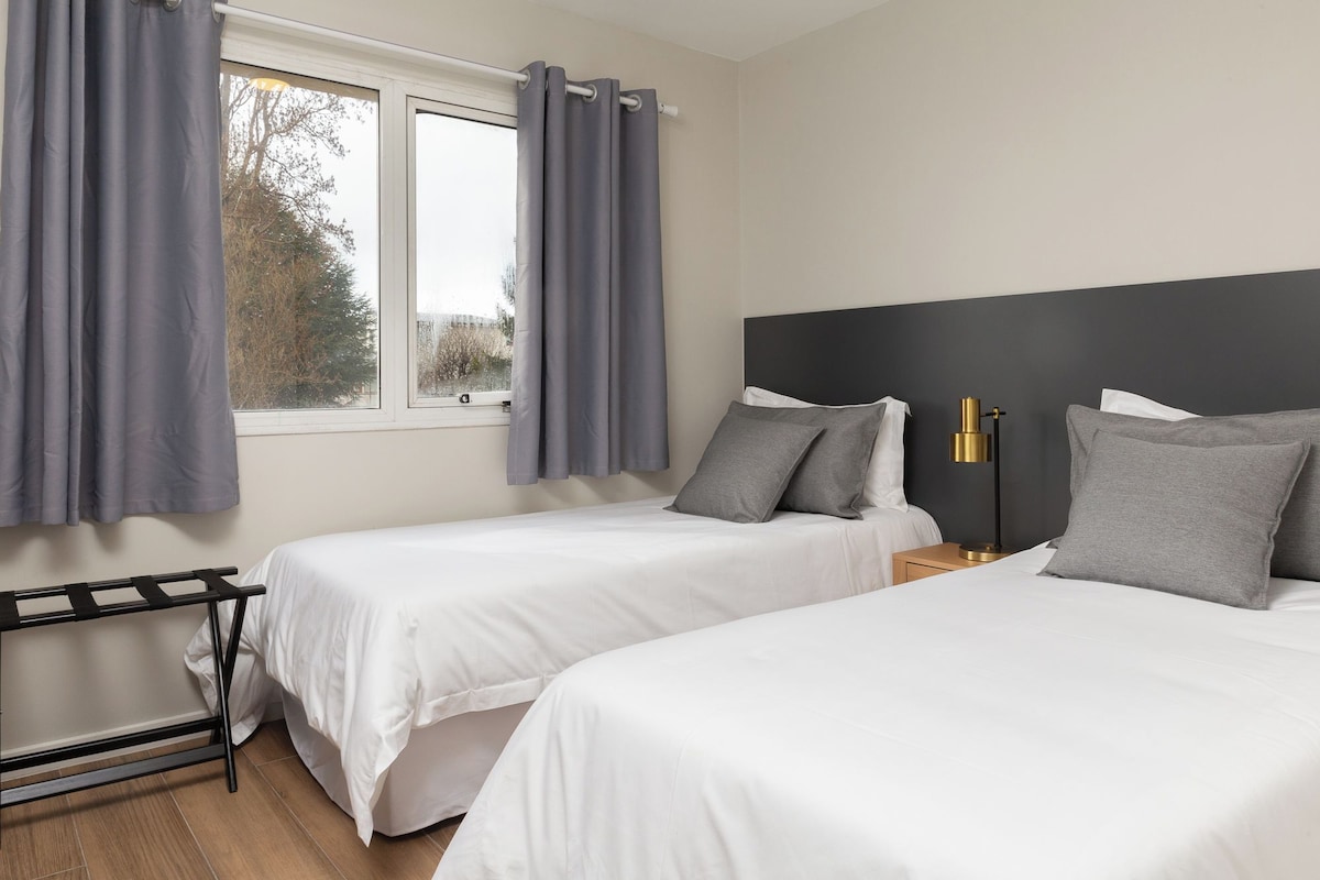 Habitación de hotel con dos camas, una ventana con cortinas, una mesita de noche con lámpara y un radiador.