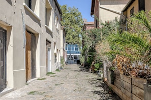 Eine enge Kopfsteinpflasterstraße, gesäumt von Gebäuden mit Wohn- und Geschäftsflächen, strahlend blauem Himmel und vielfältiger Bepflanzung.