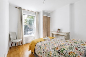 Ein Schlafzimmer mit einem Bett, einem Sessel, einem kleinen Tisch und einem Fenster mit Vorhängen.