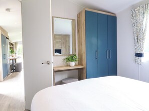 Ein Schlafzimmer mit einem Bett, einem Kleiderschrank, einem Spiegel, einem kleinen Tisch mit Pflanze und einem Fenster mit Vorhängen.