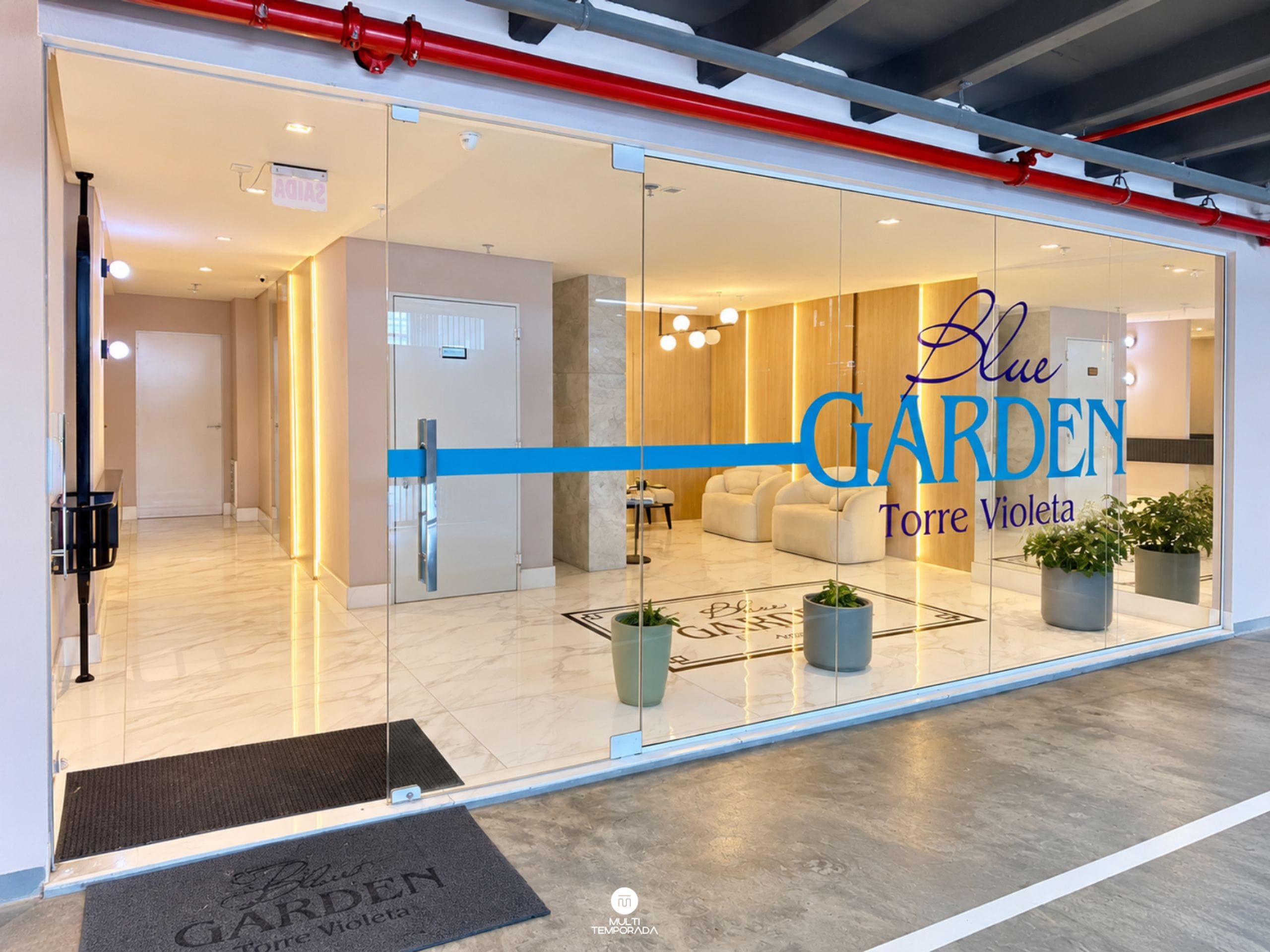 En moderne hotelindgang med en glasfacade, et receptionsområde og et siddeområde, der kan ses igennem glasset.