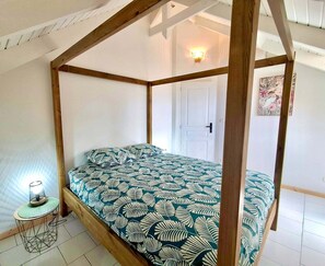 Una cama con dosel y colcha con estampado de hojas, una pequeña mesa redonda con una lámpara y una puerta al fondo.