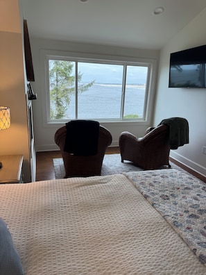 A bedroom with a large window, a bed, two chairs, and a TV on the wall.