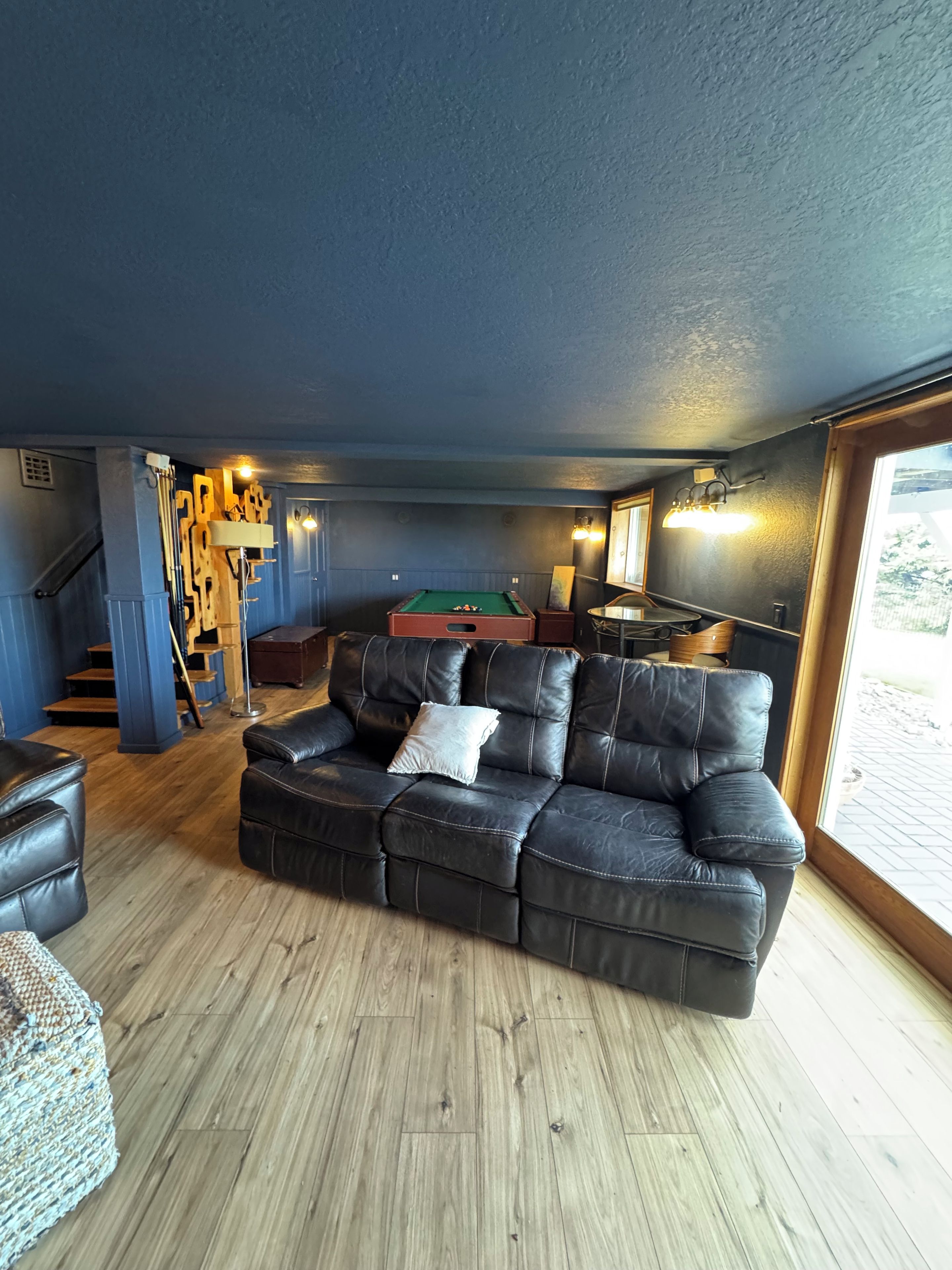 A room with a dark-colored leather sofa, a pool table, and a wooden floor.