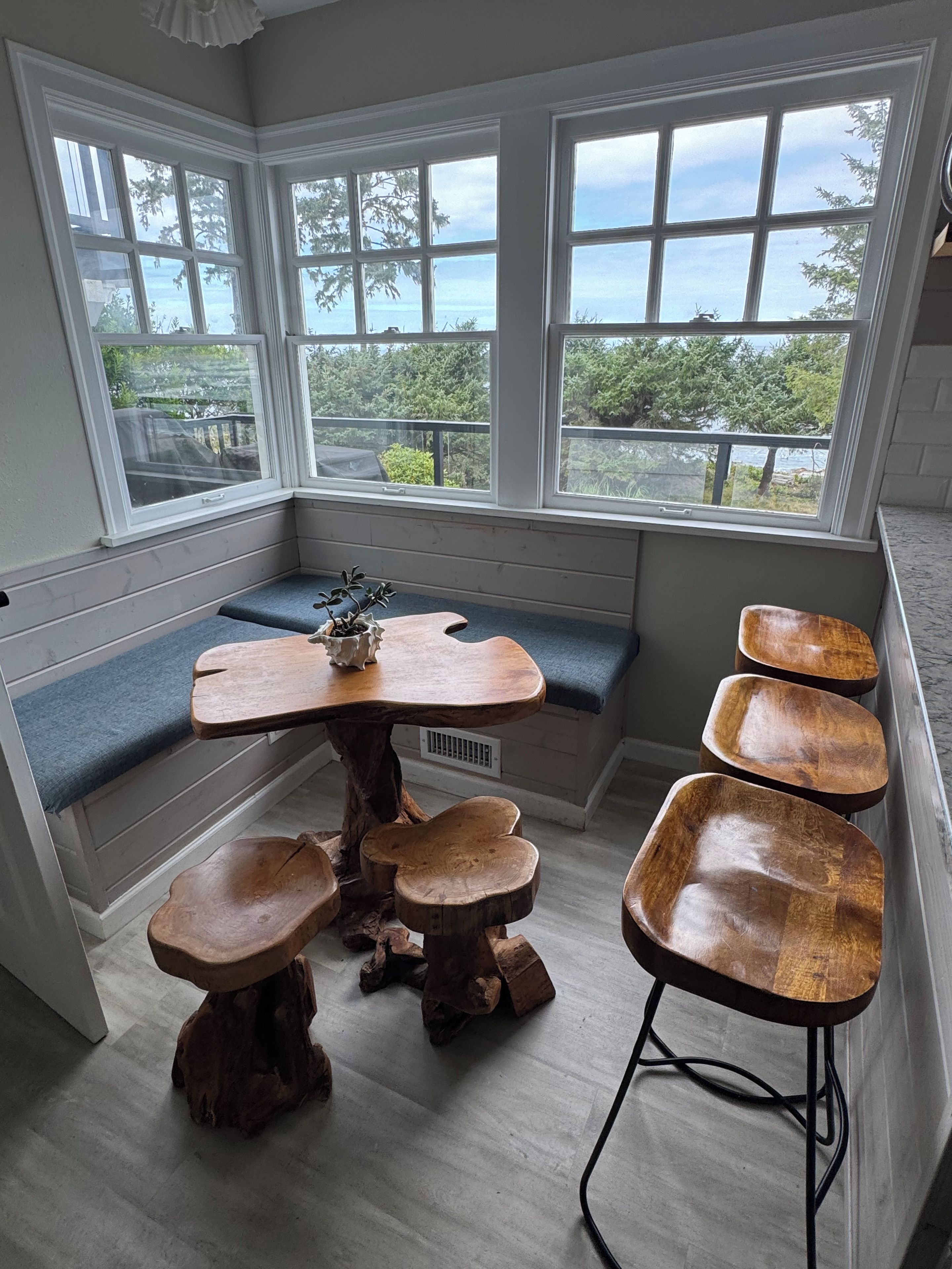 Un coin salon avec une table et des bancs en bois, de grandes fenêtres offrant une vue sur la verdure et l’eau.