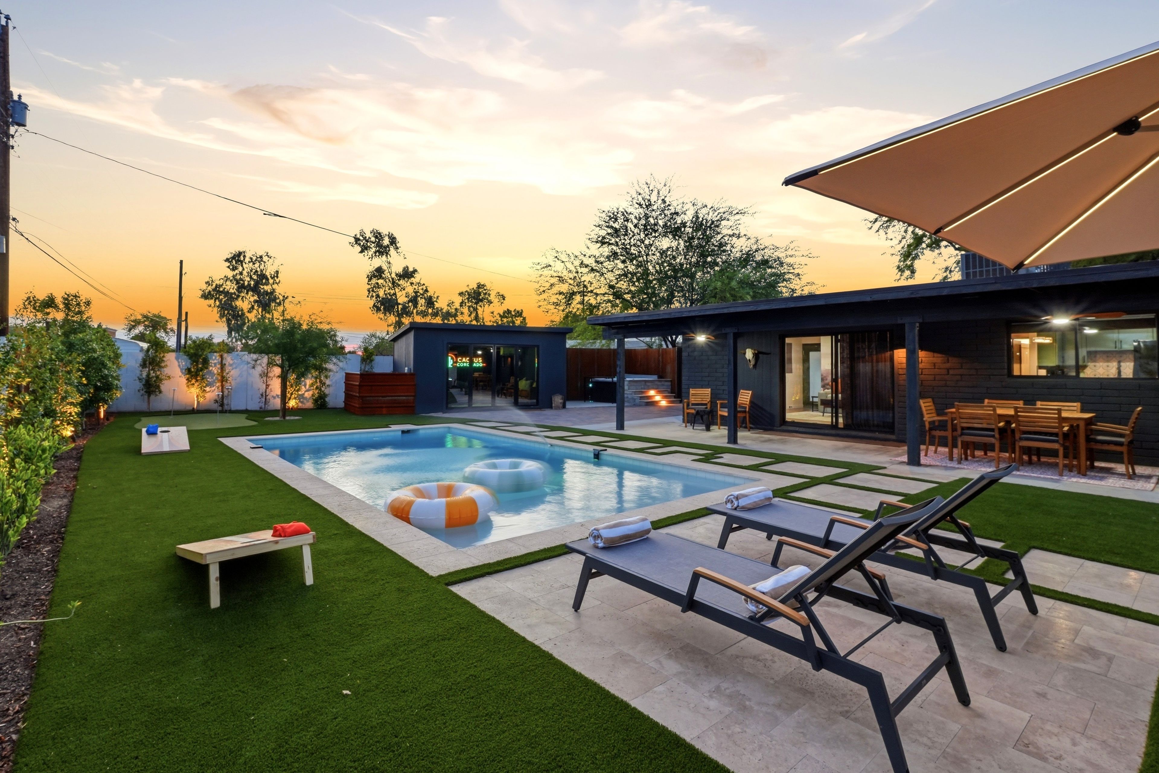 Un área de piscina moderna al aire libre con sillones, una pequeña mesa y un espacio de estar cubierto.