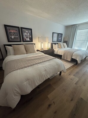 Habitación de hotel con dos camas, una mesita de noche con lámparas, revistas deportivas enmarcadas en la pared y una ventana con cortinas.