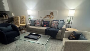 Ein modernes Wohnzimmer mit einem Glastisch, einem beigen Sofa mit gemusterten Kissen, einem dunklen Sessel und einer Stehlampe.