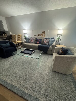 Una sala de estar moderna con una mesa de centro de vidrio, un sofá color beige con cojines estampados, un sillón oscuro y una lámpara de pie.
