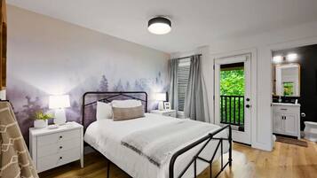 A bedroom with a bed, bedside tables, a nightstand, a lamp, a wall mural, a door leading to a balcony, and a bathroom with a mirror and cabinet.