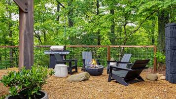 Une terrasse extérieure équipée d’un barbecue, d’un foyer et d’un espace salon, le tout entouré d’une clôture et d’arbres.