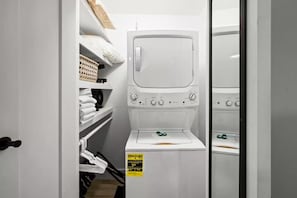 A compact laundry room with a washing machine, shelves with folded towels, and a basket.