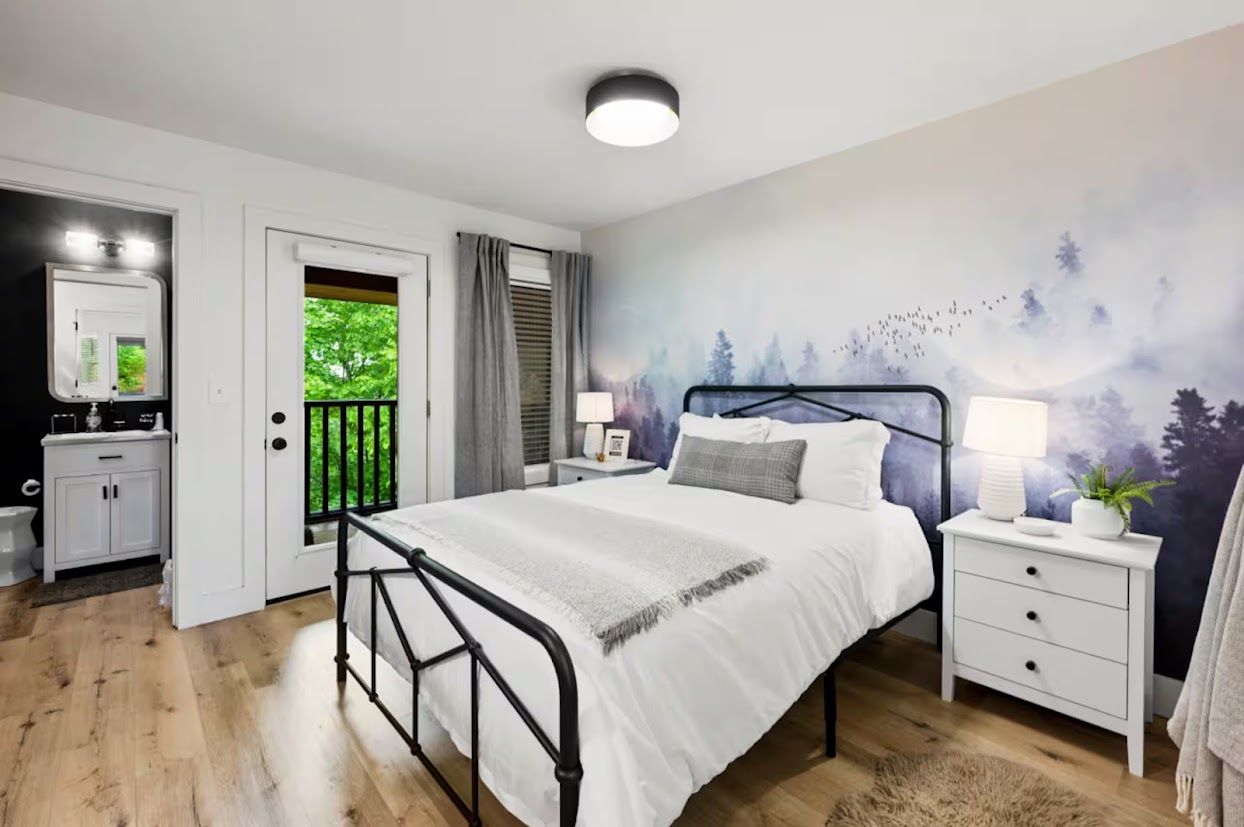 Une chambre à coucher comprenant un lit, des tables de chevet, une commode, un miroir, une porte et une vue sur la verdure à l’extérieur.