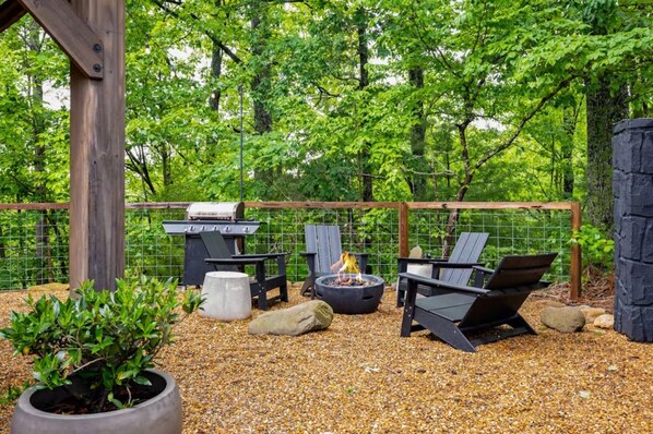 An outdoor patio with a grill, fire pit, and seating area surrounded by a fence and trees. - Pool + Arcade Resort Suite | 18 Guests (Sevierville)