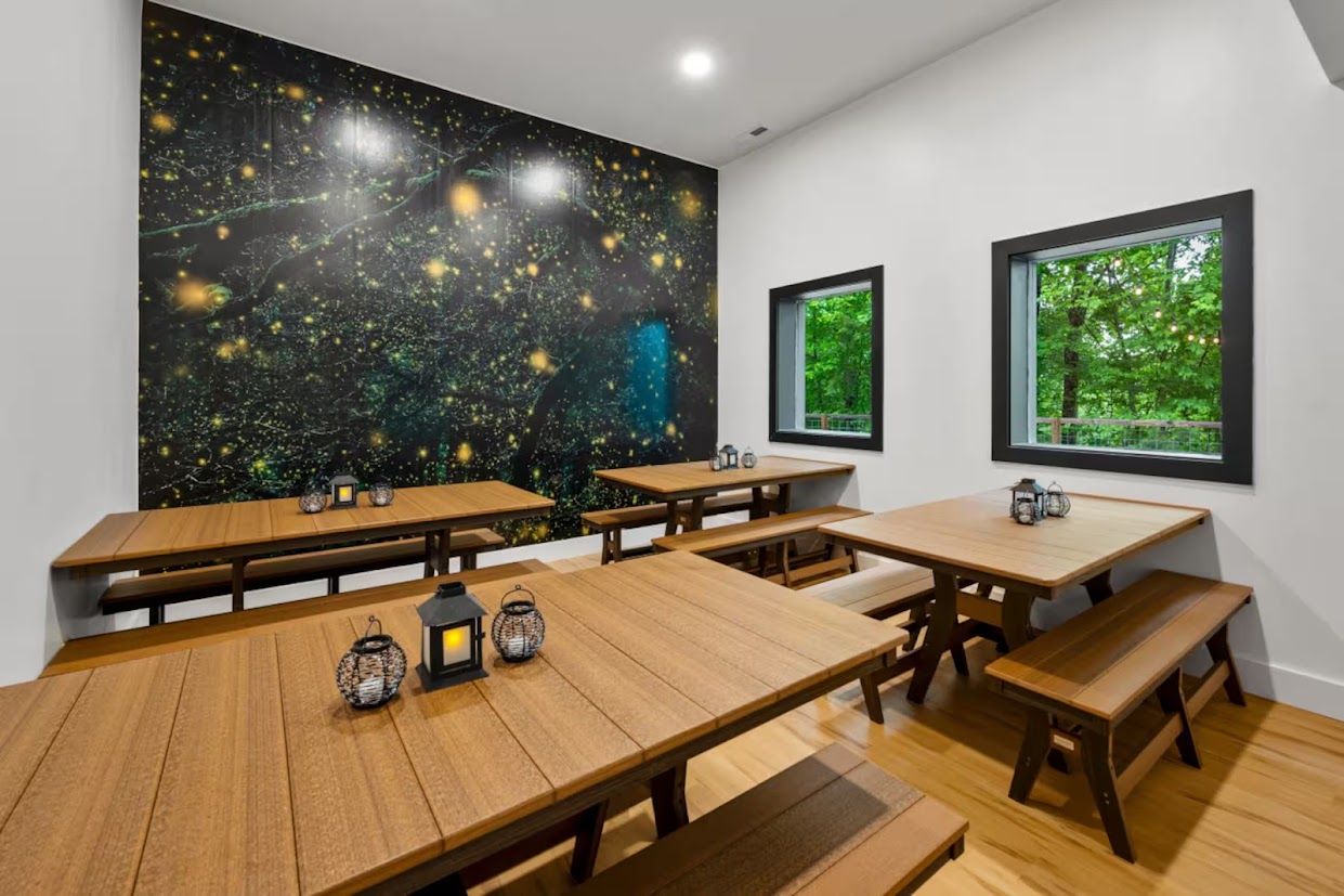 A dining area with wooden tables and benches, a starry wall mural, and windows with a view of greenery.