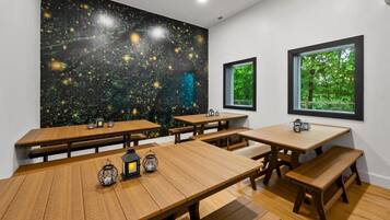 A dining area with wooden tables and benches, a starry wall mural, and windows with a view of greenery.