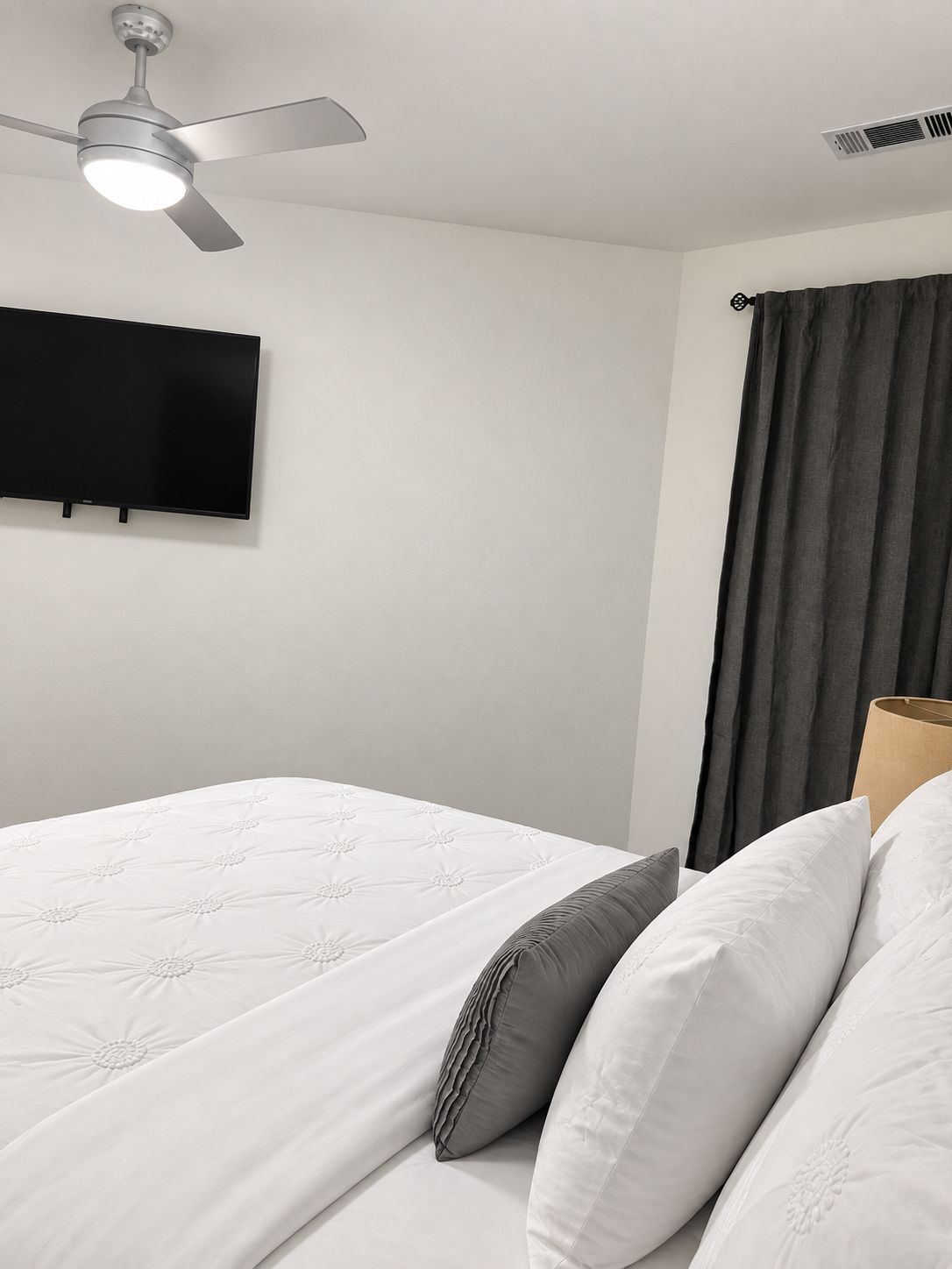 A hotel room with a bed, a ceiling fan, a flat-screen TV mounted on the wall, and a window with curtains.