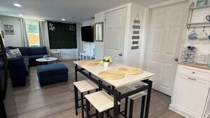Un salon moderne avec un canapé d’angle, une table à manger avec des tabourets, un téléviseur à écran plat et un mur vert agrémenté d’une ancre décorative.