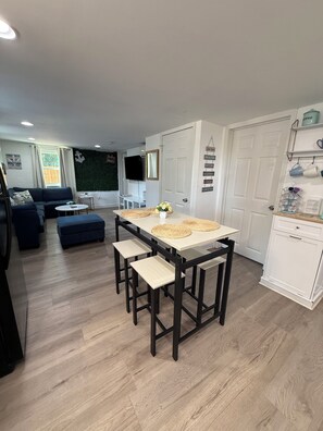 A modern living room with a sectional sofa, a dining table with stools, a flat-screen TV, and a green wall with a decorative anchor. - Cozy 2-bedroom apartment with AC in charming Mooresville (MOORESVILLE)