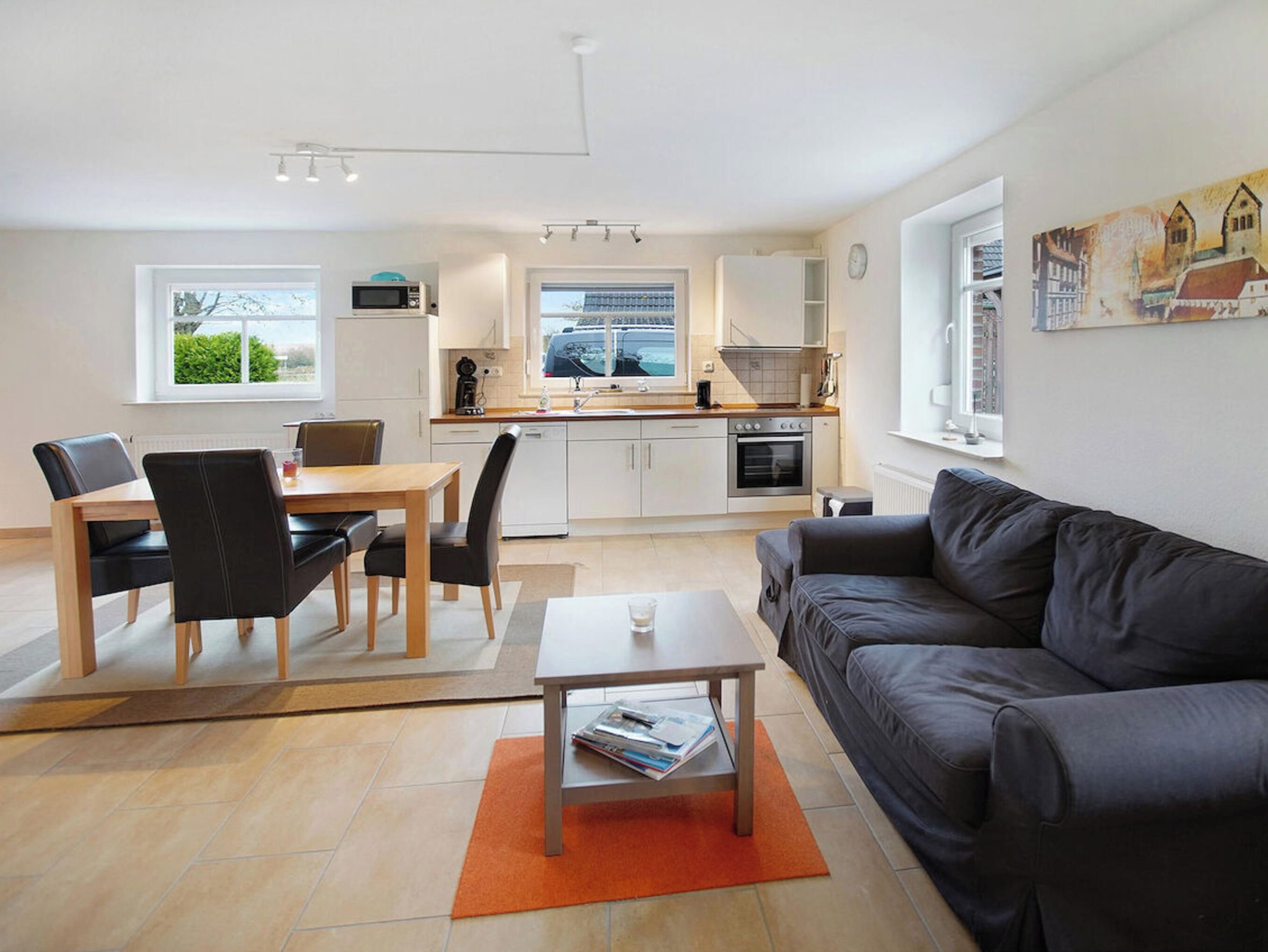A modern living room with a sofa, a dining table with chairs, a kitchen area with a microwave, oven, and sink, and a window with a view of greenery.