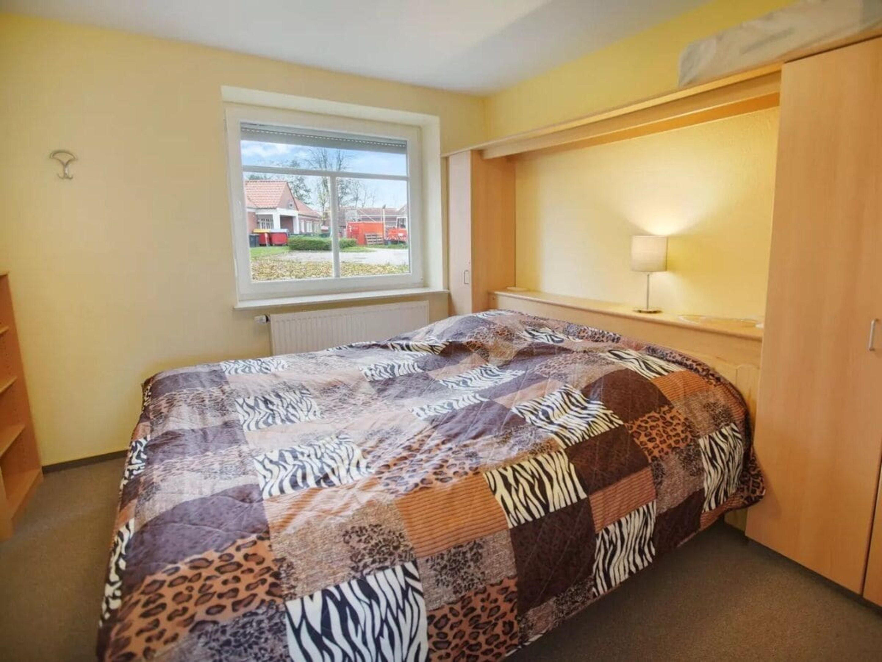 A bedroom with a zebra-patterned bedspread, a window with a view of houses, and a wooden wardrobe.