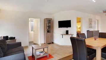 Ein modernes Wohnzimmer mit einer Couch, einem Couchtisch, einem Esstisch mit Stühlen, einem an der Wand befestigten Fernseher und einem Schrank.