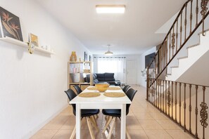 Uma sala de jantar com uma mesa posta para quatro pessoas, ladeada por cadeiras, e uma escada com grades ornamentais que leva a um andar superior.