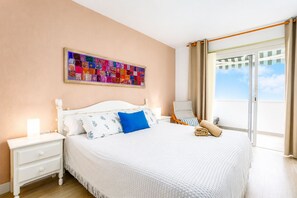 Ein Hotelzimmer mit Bett, Nachttisch, Stuhl, Fenster mit Vorhängen und einem farbenfrohen Kunstwerk an der Wand.