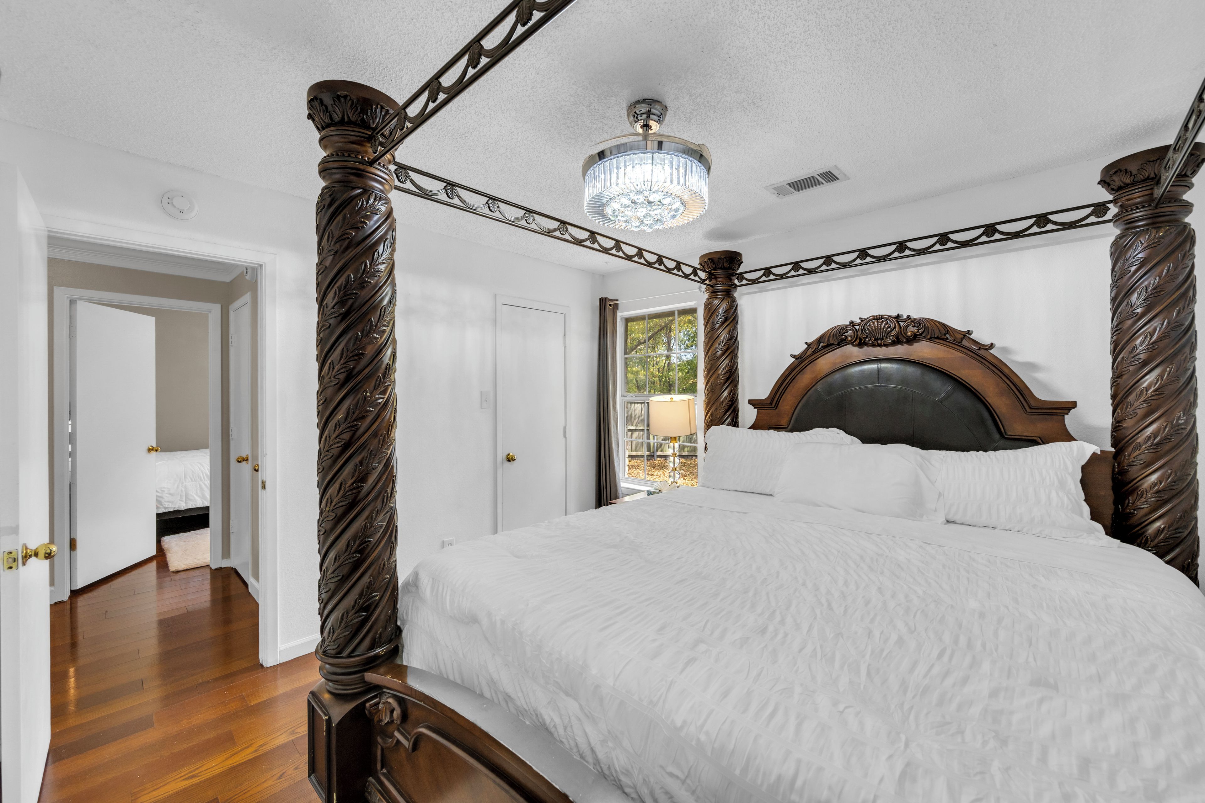 A four-poster bed with a chandelier, a wooden floor, and a door leading to another room.