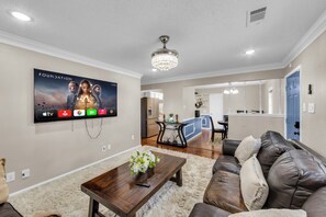 Een woonkamer met een flatscreen televisie aan de muur, een kroonluchter, een leren bank en een houten salontafel.