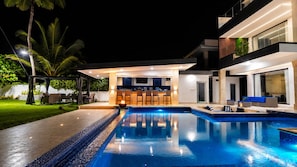 Un espace piscine extérieur moderne, doté d’un bar et de coins salon, entouré d’une terrasse bien éclairée et d’une végétation luxuriante.
