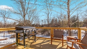 A wooden deck with a grill and chairs, overlooking a snowy landscape.