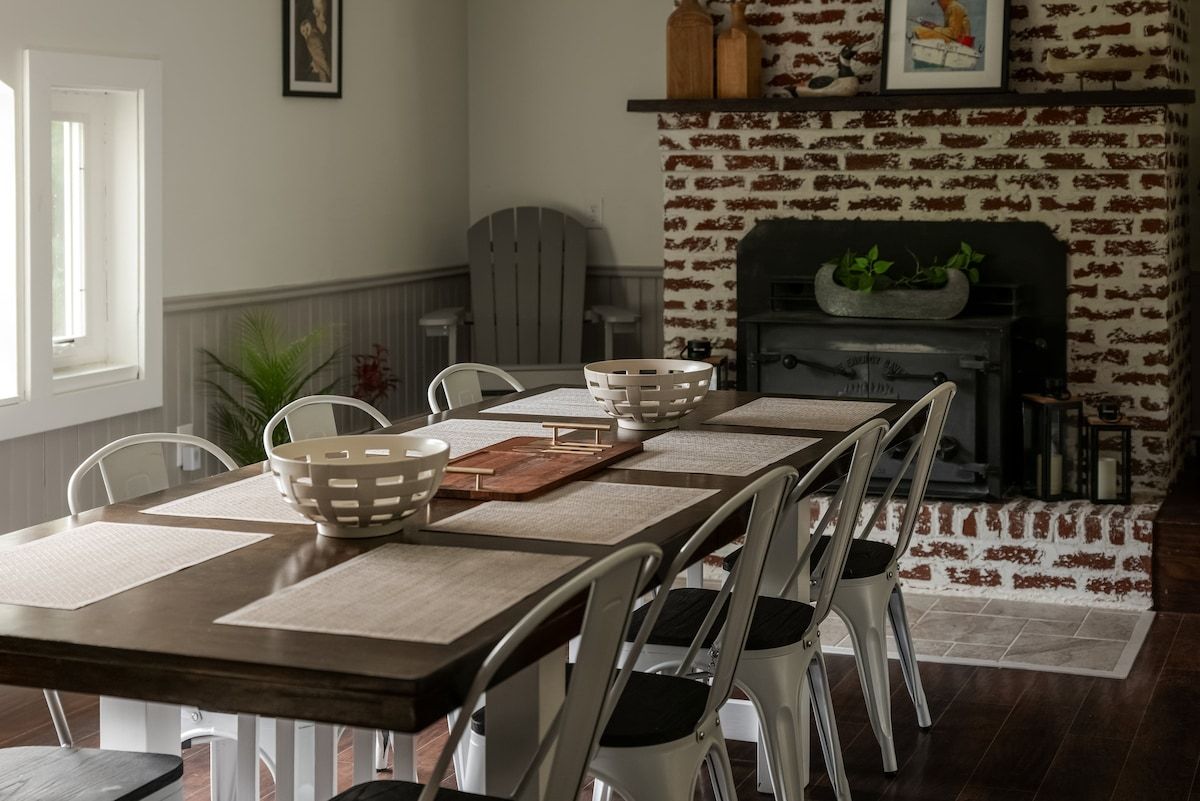 A dining area with a long table, white chairs, and a brick fireplace.
