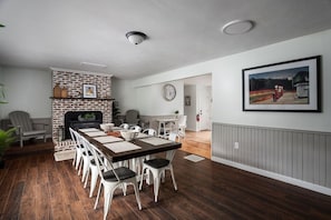 Een eetruimte met een stenen open haard, witte stoelen, een lange houten tafel en een klok aan de muur.