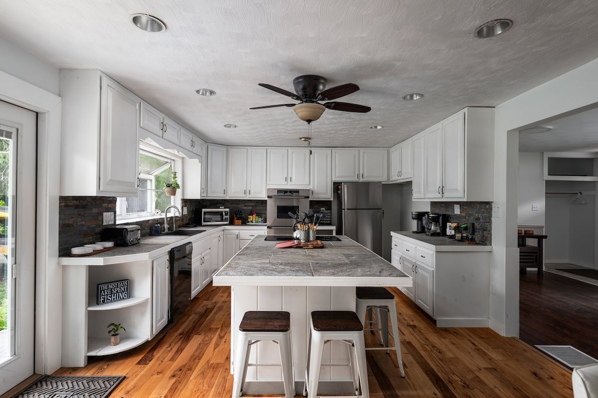 A modern kitchen with white cabinetry, a central island with stools, a ceiling fan, and a view to the outdoors.