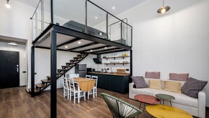 Een modern interieur met een trap naar een vide, een eethoek met tafel en stoelen, een keuken met een ingebouwd kookeiland, en een zithoek met een bank en bijzettafels.