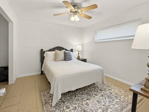 A bedroom with a bed, a nightstand, a lamp, a ceiling fan, a window with blinds, and a patterned rug.
