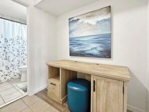 A bathroom with a shower curtain, a toilet, a wooden vanity with a stool, and a painting on the wall.