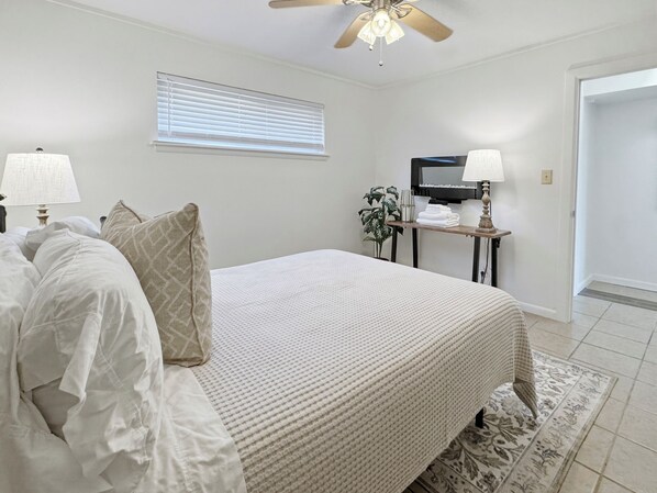 A bedroom with a bed, bedside lamps, a ceiling fan, a window with blinds, a bedside table with a lamp, and a plant.